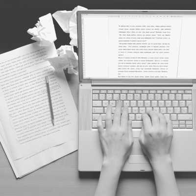 Two arms are typing on a laptop, with crumpled pieces of paper lying about.